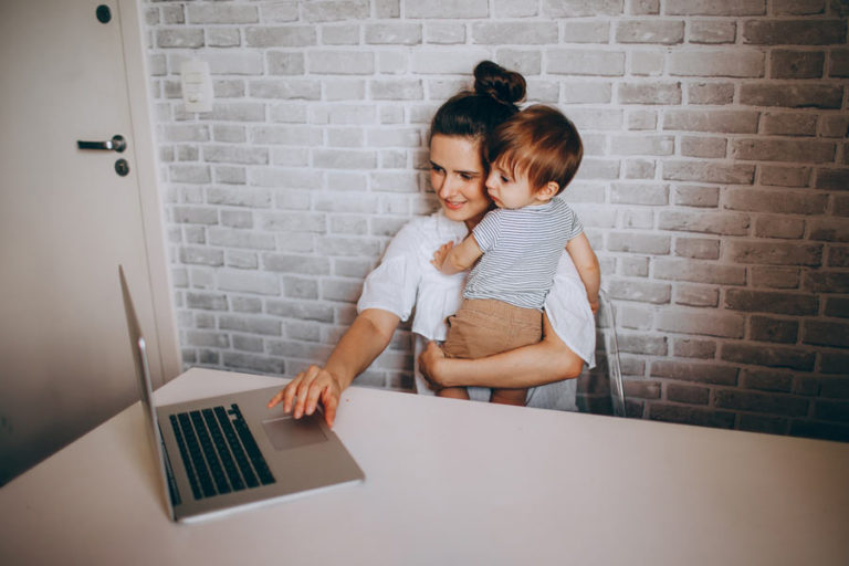 mom holding toddler while using her laptop in front of the backdrop of a whitewashed brick wall.
